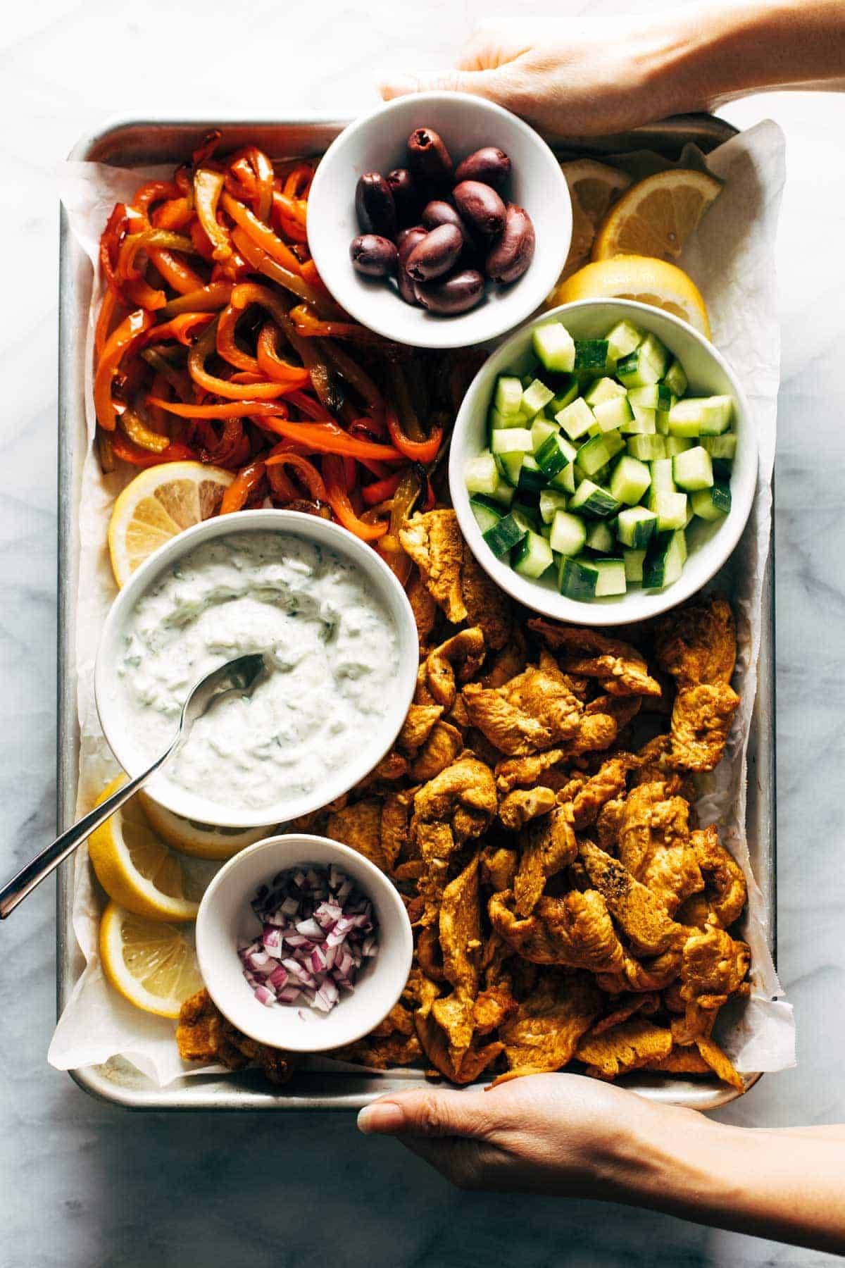 Chicken and peppers on a sheet pan with olives, cucumbers, and tzatziki.